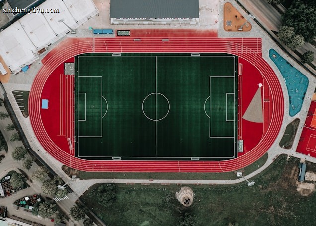 An aerial view of a large sports complex featuring a central green football field surrounded by a red running track. Adjacent to the track, there is a parking area, buildings, and pathways. Nearby, there are additional sports courts in different colors, including a blue swimming pool and orange basketball courts.