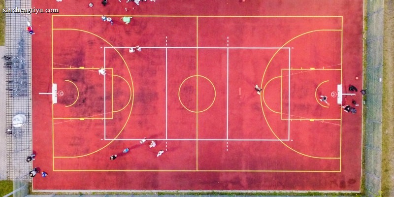 An aerial view of a sports court with a vibrant red surface and yellow lines marking different sections for basketball and soccer. Several people are present on the court, some engaged in activities, while others are standing along the perimeter. The court is enclosed by a metal fence and surrounded by grass on all sides.